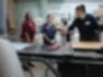 A person named Ava demonstrates woodworking in a workshop. Others watch attentively. Tools and machinery seen in the industrial setting.