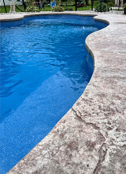 The side of an in-ground swimming pool with curved edges and a stamped concrete pool deck.