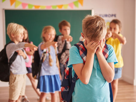 Crianças em sala de aula apontando na direção de um colega que está chorando e escondendo o rosto, mostrando um momento de bullying na escola