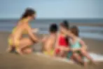 Família de quatro pessoas na praia, com duas crianças e duas adultos, desfrutando de um dia ensolarado na areia, sorrindo e interagindo juntos.