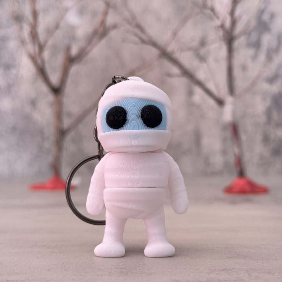 Adorable 3D printed mummy keychain figure with blue face and black eyes, posed in front of a soft-focus wintery backdrop.