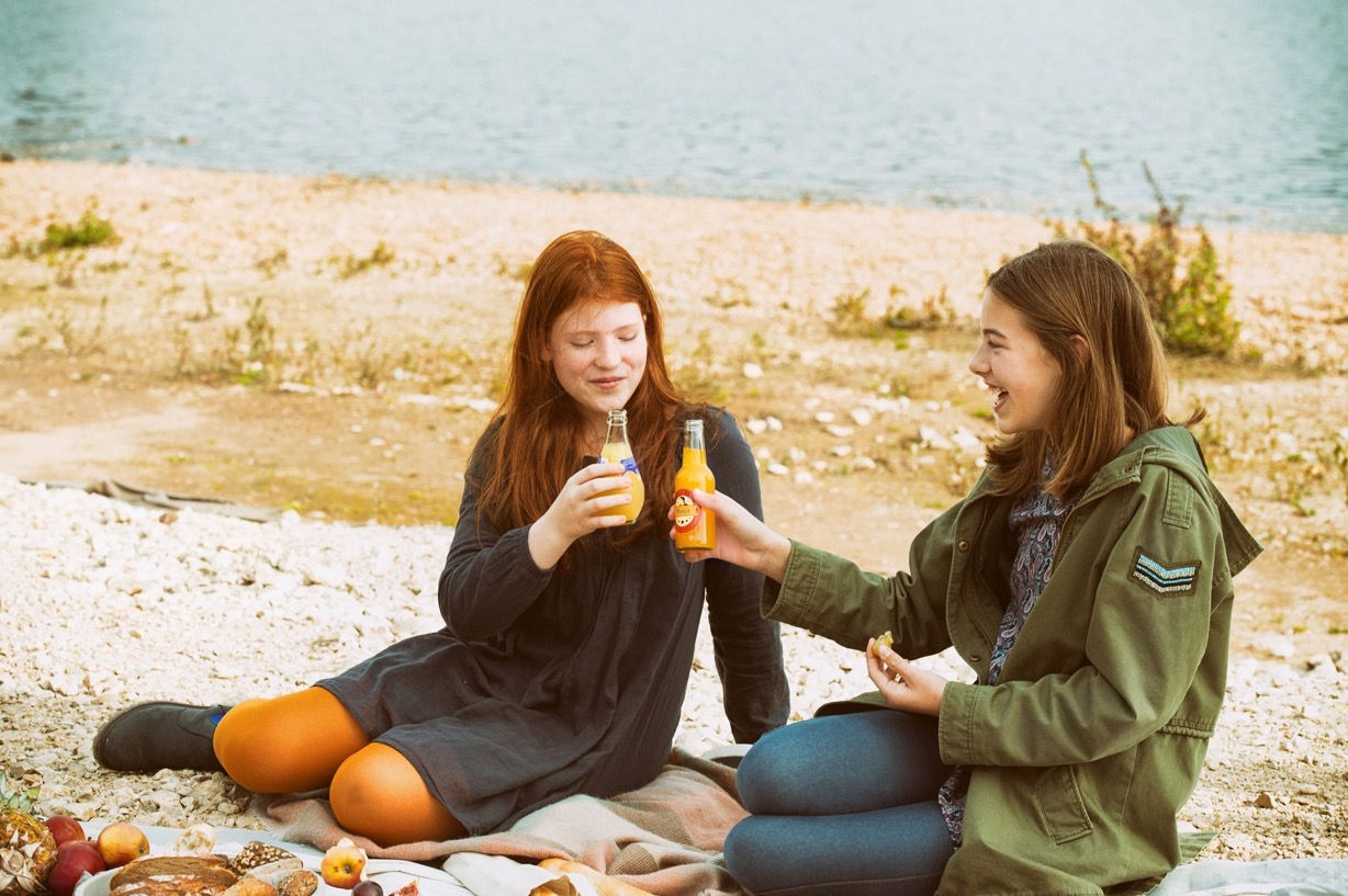 Freundinnen genießen Getränke am See – entspannte Outdoor-Werbefotografie für Health & Wellness Marken.