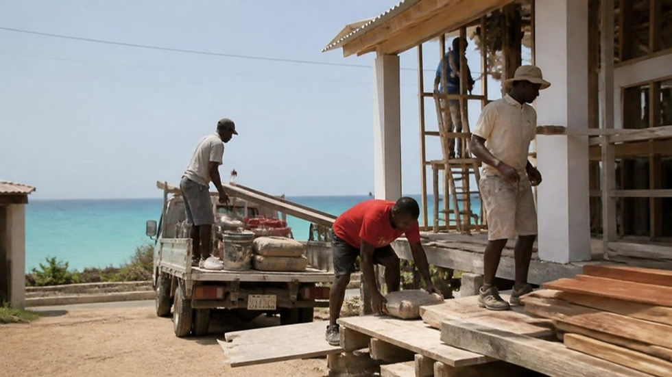 Obreros raizales construyen una vivienda en Providencia mientras descargan materiales de un camión frente al mar Caribe.