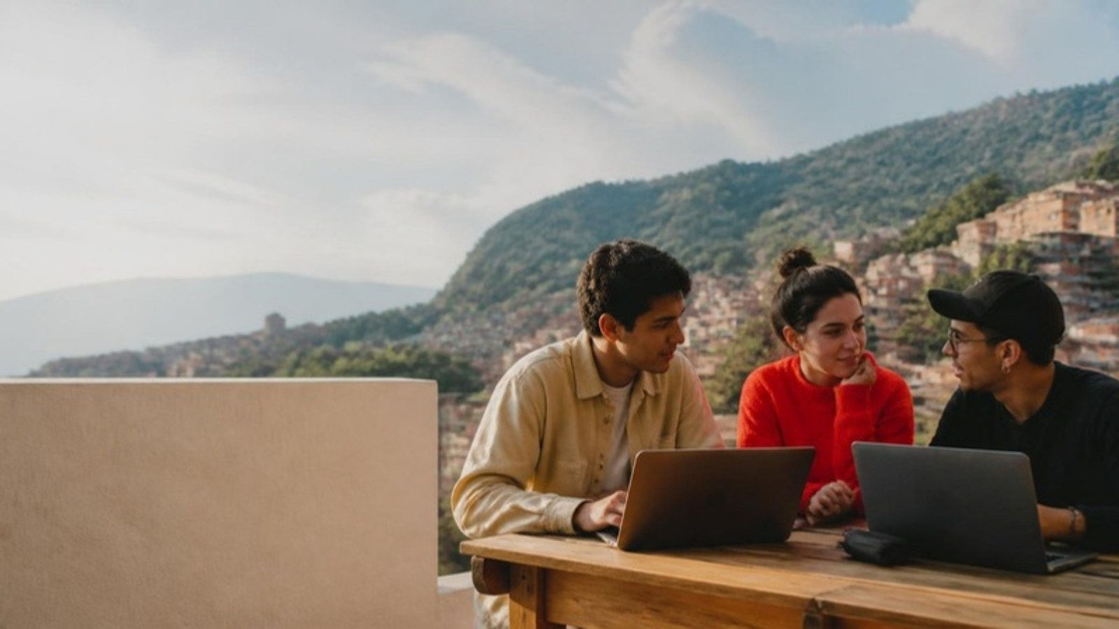 Jóvenes emprendedores trabajan en portátiles en una terraza con vista a las laderas de ladrillo de Medellín.