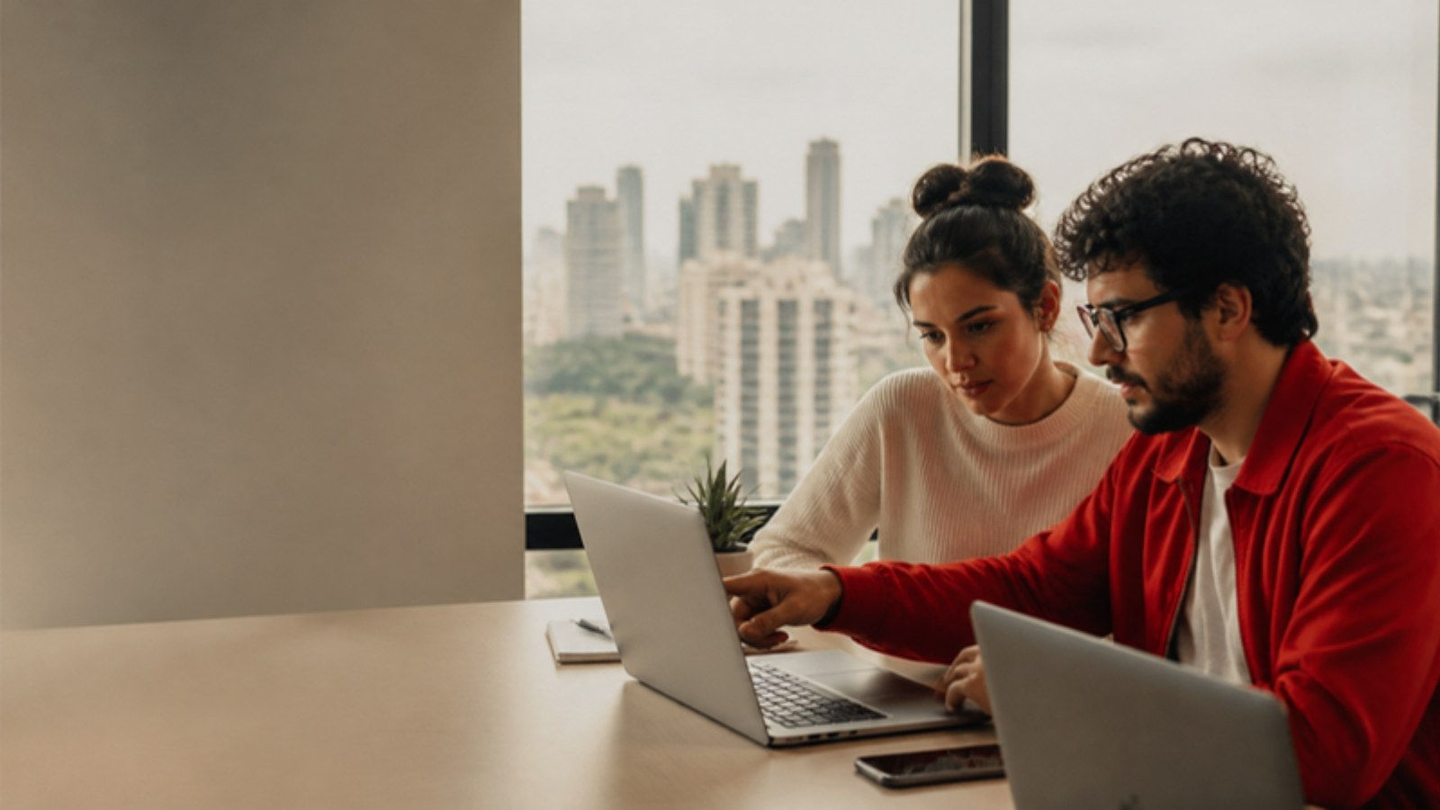 Dos fundadores de una startup trabajan con portátiles en una oficina de São Paulo con la ciudad al fondo.