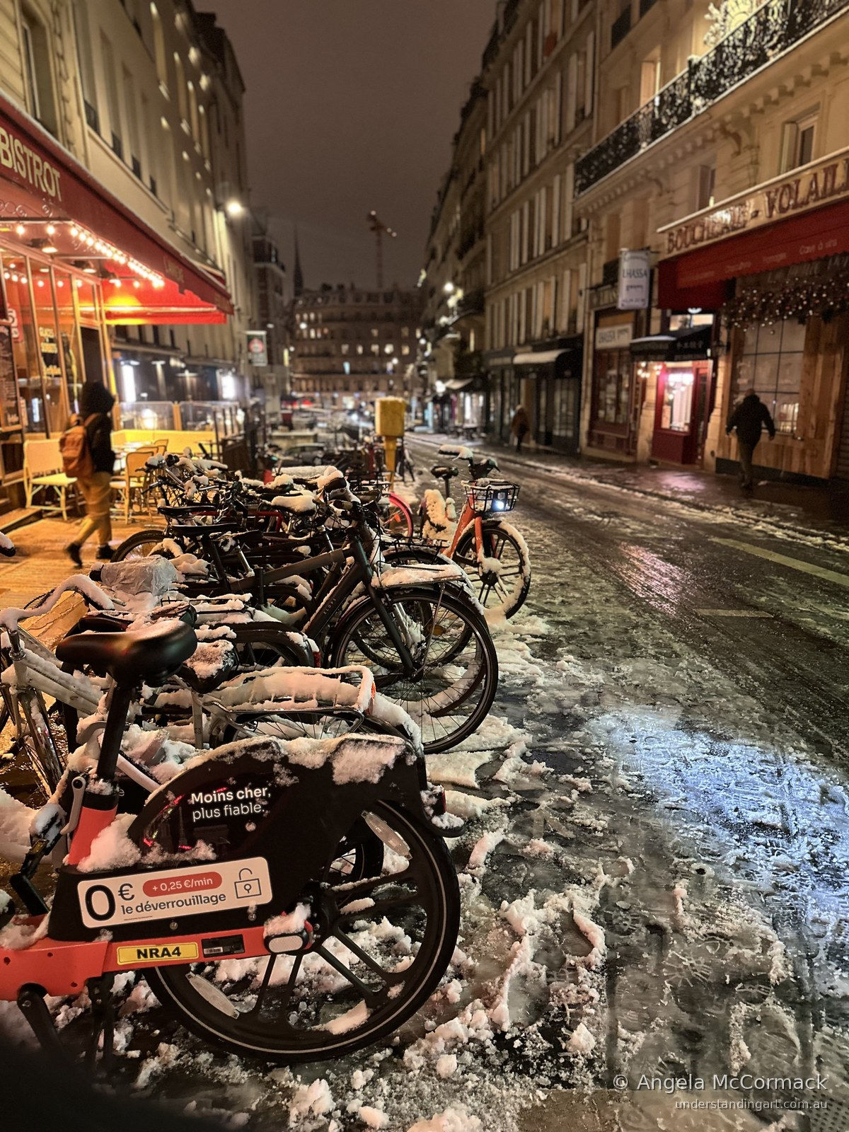 Snow-covered bicycles lined up outside a lit bistrot on a Paris street at night