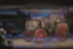 Historische schwarze Kutsche mit roten Speichenrädern in dunklem Museum/Depot, neben Oldtimern, unter Stellplätzen 7 und 8.