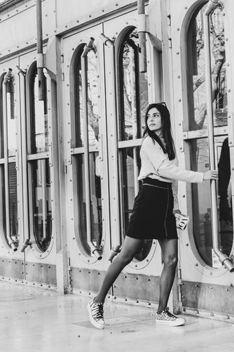 Portrait en noir et blanc d'une jeune femme en minijupe et baskets, posant de manière dynamique devant les grandes verrières métalliques de l'Eden Théâtre à La Ciotat, style street photography.