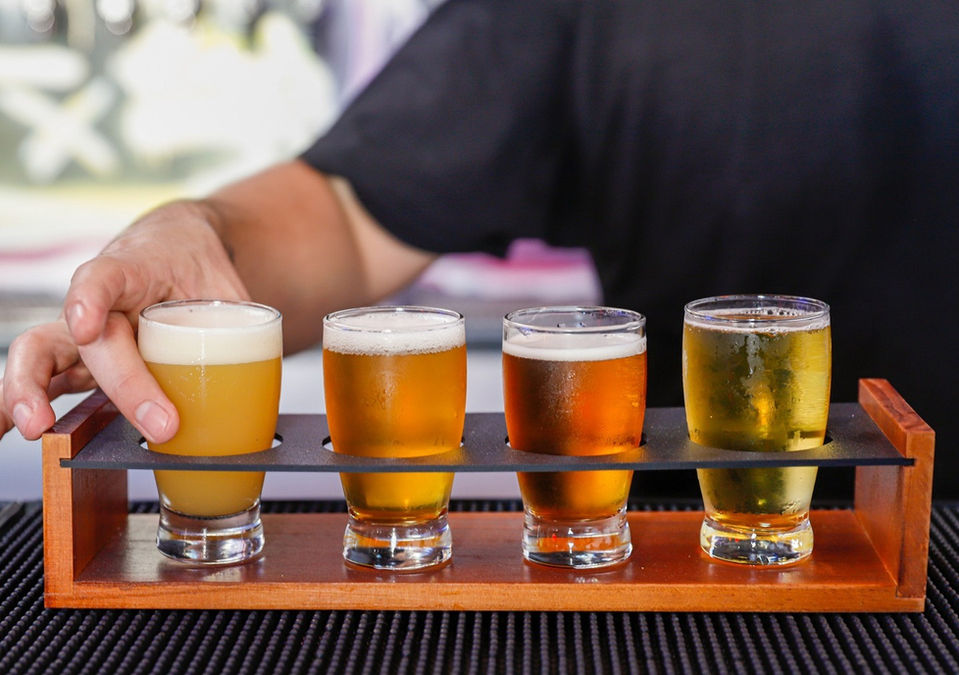 A bartender’s hand placing the last of four small glasses into a wood & glass flight rack, a NICE one! Sitting atop a black barmat, his arm and black Tshirt visible but way blurry in the background. Each beer a distinctively different color… mmmmm