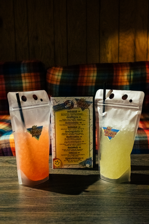 another view of the playing card box, upright this time, standing betwixt two standing cocktail baggies, iced with straws, one orange, one yellow, dark background at My Friend's House in RPM