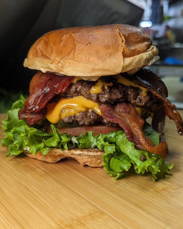 Sideview of a large overstuffed double-cheeseburger with huge slices of bacon hanging out, again like tongues, all on a toasted bun, atop a wood-grained surface, blurred background.