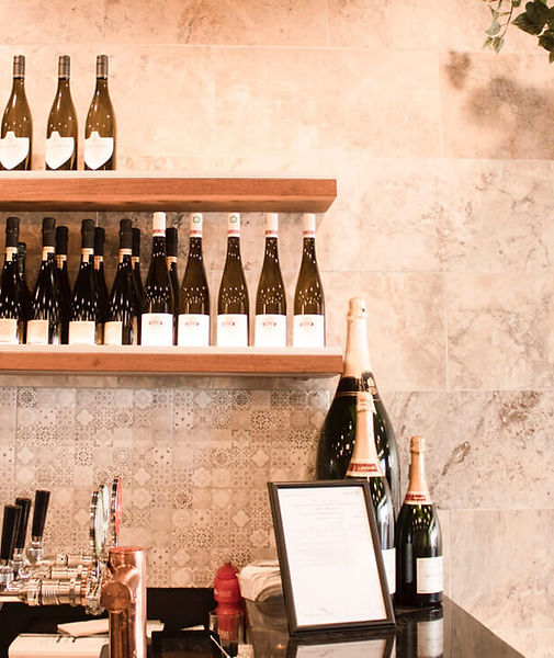 a curiously composed shot of a shallow wine rack and top fo a bar counter with 3 champagne bottles and 3 taps and various notecard items, against a rough plaster wall with a sprig of tree leaves entering top-right