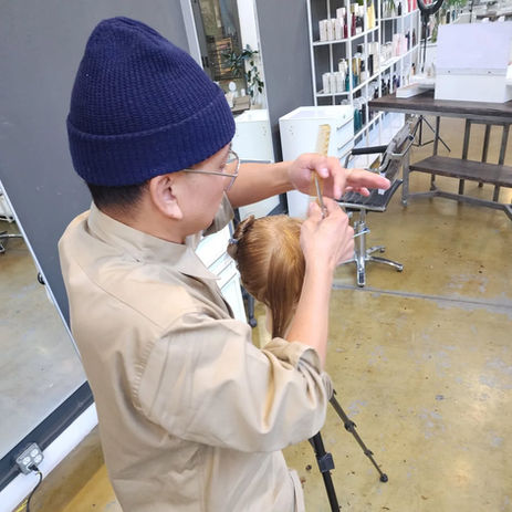 Nathan Nguyen, seen hear from his above-right, working the hair on his demo mannequin with both hands, big blue beanie now more prominent, floor and product shelves in background.