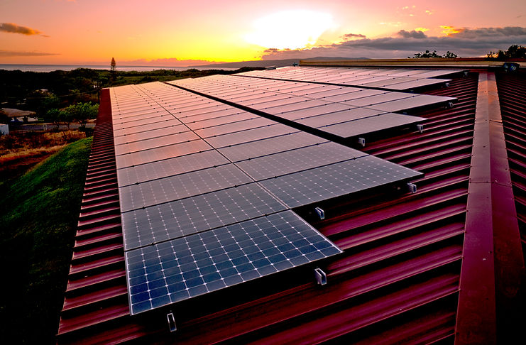 a dramatic sunset shot of a metal roof with extensive solar panels installed, stretching into the distance and trees and a bay beyond to the horizon with a golden sunset