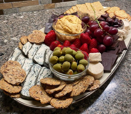 An oval charcuterie featuring slices of bleu cheese, whole grain crisps, pimento olives, hummus, strawberries, grapes, chocolate and black olives crowding a large oval plate on a hard granite surface.