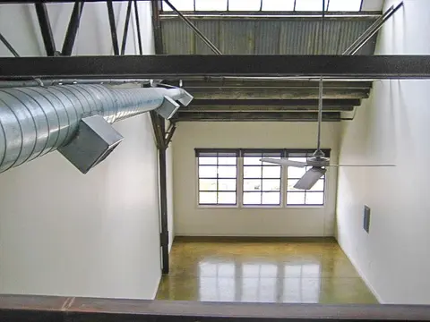 a tight view of the ceiling & silver duct work in the upper area of a work/live loft in the Claremont Packing House with hazy sunshine pouring in from above skylights and loft railing in brown metal and various elements of an open-rafter ceiling.