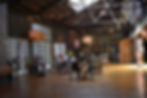 a quiet indoor atrium setting in the Claremont Packing House with medium skylighting, lots of wood surfaces & browns, a few people sitting at small round tables with lots of space around them and the entrance glass-paned doors to Augie's Coffee House visible along the right side wall