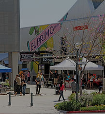 a setback shot of the Claremont Packing House public square next to the parking structure, with pedestrians, popup shades, and the east wall of the CPH with gaily painted graphics and small trees and 2 bollards