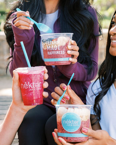 five womens' hands holding three Nekter items, two are wide clear-plastic fruit cups and one is a short clear-plastic fruit drink, can just see the front of one woman's smiling face on the right, and another woman's body in the center, and another woman's hand reaching in from the left side