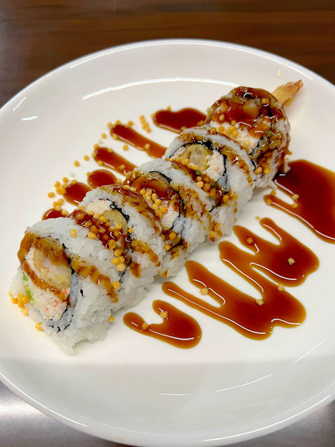 An overhead view of a medium-size white ceramic plate of five crab rolls, with sesame seeds, seaweed, ginger and heavy drizzle of thick dark brown sauce, decoratively squiggled.
