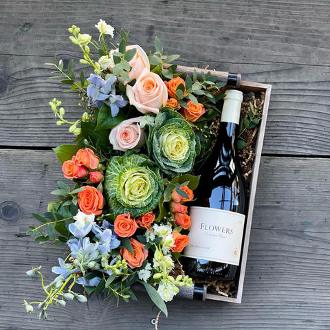 looking straight down upon a more Spring-themed flat floral arrangement in a gift box beside a fine looking bottle of red wine, lying flat down with the flowers, all atop a gray wooden slatted surface, quite beautiful