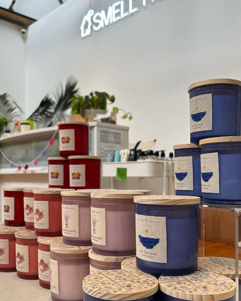an angle-on view of various stacks of jar candles in basic colors with labels that remind of jams, shelves stretching into the background where we see part of a white neon Smell House internal sign high up on the wall