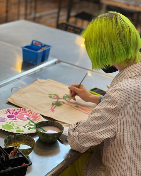 a woman with neck-length straigh green hair working with a brush doind watercolor on paper at a worktable, with bowl of murky water and paper plate and other paraphernalia, we can't see her face, she is wearing a loose button-down collared pinstriped shirt and a mask we can barely see