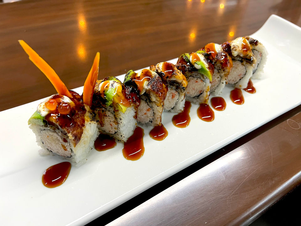 A caterpillar of crab-filled sushi rolls with two carrot antennas or ears, on a long white porcelain serving plate, drizzled with a thick soy sauce, all on a wooden tabletop.