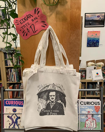 unbleached cotton Edgar Allen Tote Bags with screenprinted image of Edgar Allen Poe holding an umbrella in the rain, hanging from a nail on the side of a wooden bookshelf, with a handwritten sign that says "$12 each", book racks and wall art can be seen in the background
