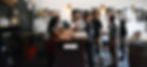 interior casual photo of a social-mixer event inside one of the vendor's spaces, with a woman at a bar serving coffee & beer to an eager line of young men, all are chatting amiably, with medium-size portrait paintings on the white wall behind