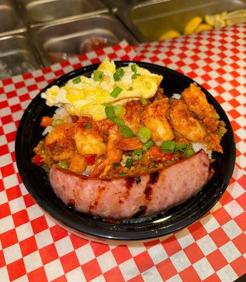 An overhead vies of a black plastic plate of a Hawaiian-style breakfast, featuring a slice of grilled Spam, scrambled egg, spicy row of shrimp with green onions and white rice, all on tablecloth of red & white checkered paper and blurry serving trays visible top background.