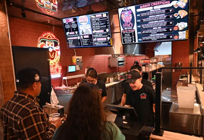 Typical scene at the Fred's Red Taco order window of their booth at RPM, with three employees taking orders, two at small register screens, and the two bigscreen menus filled with infos above. The kitchen areas can be seen behind, with two more food preparers working at their stations