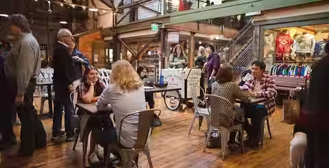 a casual scene of the central atrium area with small tables spread about and peopled with happy strollers and diners and coffee drinkers, staircase and storefront structures visible behind