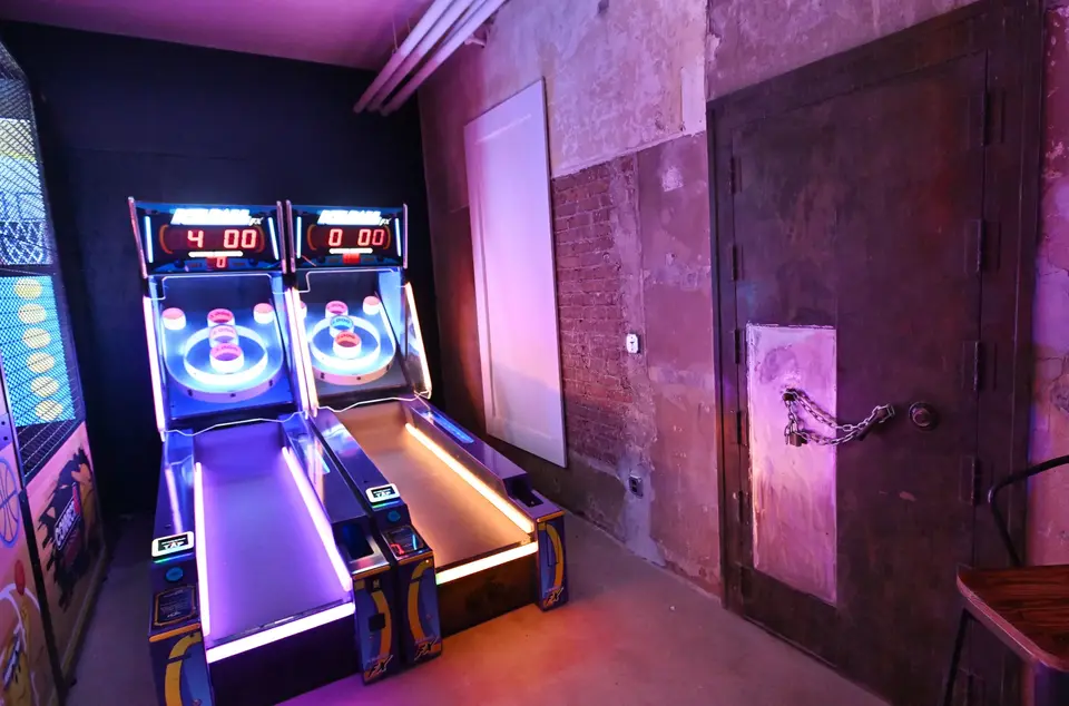 A no-person shot of the back corner of the Superslice Arcade featuring two side-by-side bowling-alley cornhole consoles, all lit up with neon lights and the bare brick and concrete industrial architecture of RPM with a big metal door, closed and locked with a heavy chain… all very atmospheric.