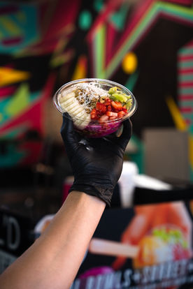 A guy’s hand holding a freshly-cut fruit clear plastic cup out away from the camera, black safety gloved, forearm showing, all against a background of edgy pop blurry shapes and dark blacks, very cool and painterly composition overall.