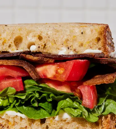 A very much closeup of a sliced bacon, lettuce, and tomato sandwich on whole wheat bread with plenty of fresh green lettuce and a healthy amount of mayo barely oozing top and bottom of ingredients, blurry white tile wall behind.