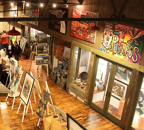 a lookdown view of an art exhibit in the central lobby atrium of the Claremont Packing House, with vintage wooden flooring and standing easels, with two visible storefronts with large glass doors & windows, and some cross girders in shadow and two people standing almost off-frame