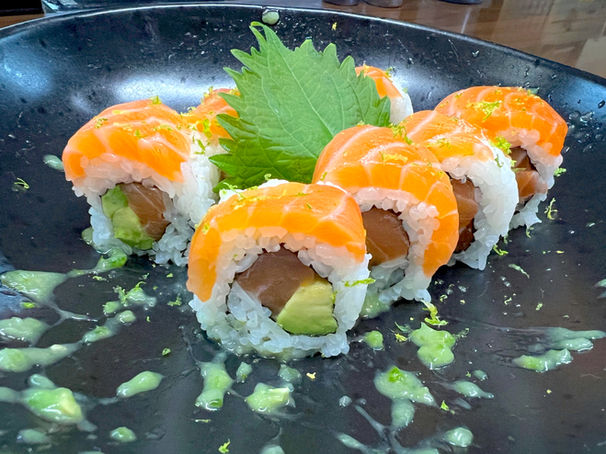 A group of seven salmon rolls with white rice and avocado and tuna in the center, all on a black plate in closeup, with splatters of wasabi splashed about, peek of wooden tabletop just behind.