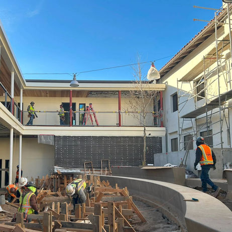 The inner-dorm mini-amphitheatrical greenspace takes shape as several construction guys move about working on the forms for a complex concrete pour, hard hats and reflective vests, ladders, hanging lamps, blue sky, white walls and scaffolding.