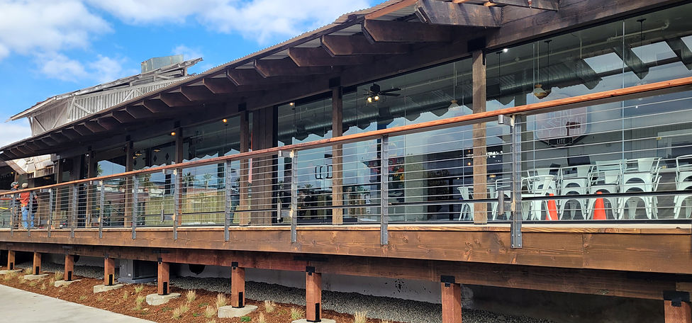 A daytime longview of the very clean wooden western deck at RPM, with floor-to-ceiling windows in front of the shops and sophisticated open wire & wood guard-fencing and short wooden pilings underneath by the sidewalk, with small xeriscapic plants visible. Blue sky with clouds.