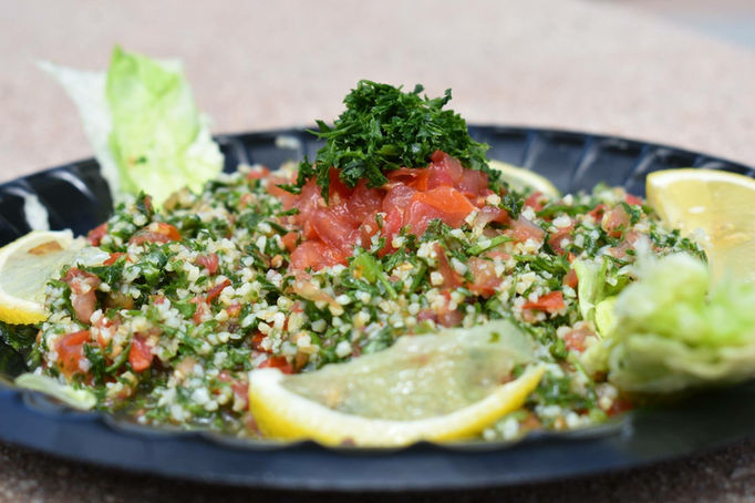 Closeup sideview of a black plastic plate full of tabouli with garnish of chopped tomatoes and basil, lemon wedges and lettuce leaves on a hard stone tabletop