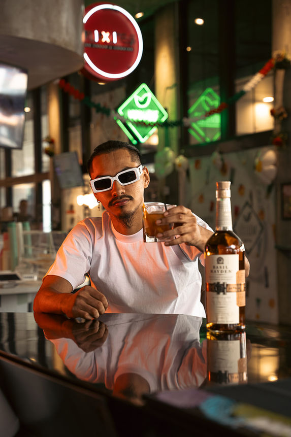 Hip young man in white shirt and big white-rimmed sunglasses sits at the Brass Monkey Social bar toasting the camera