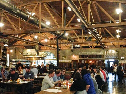a cavernous restaurant interior space with high open-rafter ceiling, drop lighting, windows and skyligts with numerous diners sitting at long wooden tables drinking beer and eating