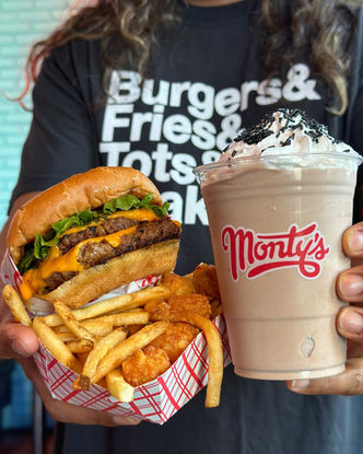 A woman’s torso in black Tshirt with the words “Burgers & Fries & Tots & (maybe) Shakes(?)” in big white sans-serif letters, we only see a bit of her hair hanging down, and fingertips holding out a big chocolate shake with whipped cream and dark sprinkles on top, then a double-cheeseburger wrapped tightly in paper, squeezed together with a paper tray of french fries.