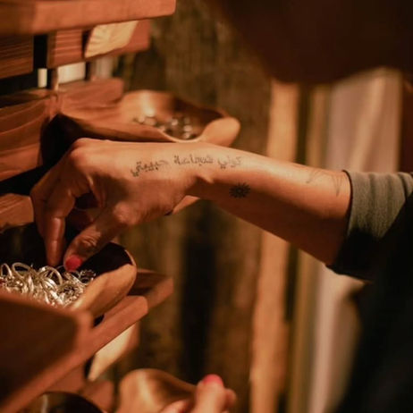 a tatooed hand picking out a hoop from a small wooden bin-shelf, on a rack with other bins, close in so we only see the forearm & hand, with blurred curtain-like elements in background