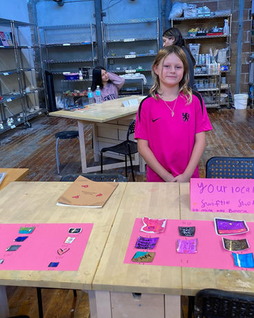 young girl in bright pink T poses behind a worktable with various cutout drawings and colorings displayed upon it, two adults can be seen right behind her, then racks of paints and equipment and drying crafts or sculptures, polished wooden floor all round underneath