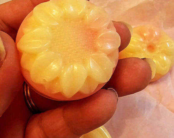 woman's fingers & thumb holding up a smallish round floral yellow soap with a circular leaf-petal design, others barely visible underneath in background on cloth