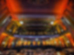 the main hall viewed from the stage with wooden chairs and tables with black tablecloths set up on main floor for a banquet and yellow gold red lights througout up to the balcony