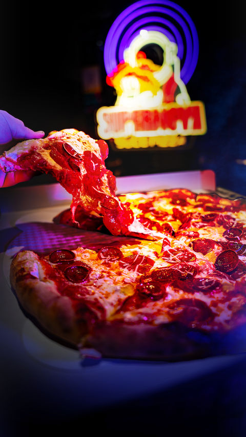 Vertical phone shot heavily processed, dramatically running to black top and bottom, a freshly cooked pepperoni pizza in a special custom opened box and the new neon Supernova Slice Pizza logo visible behind, but now very blurry. A hand is pulling a heavily-laden slice from the pie, with is in dramatic closeup and harsh lighting.