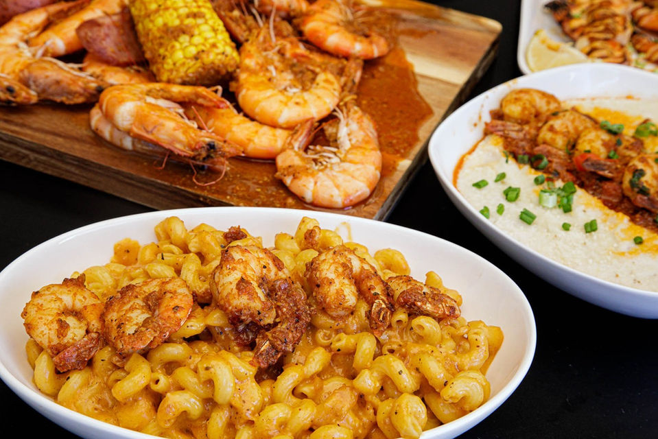Professional product shot of a group of freshly cooked dishes, with a wooden cutting board of jumbo shrimp and corn on the cob, all covered with a light bbq sauce, spicy shrimp and mac & cheese, a bowl of spicy shrimp over jasmine rice, and just a wee corner of blurry grilled shrimp with lemon wedge.